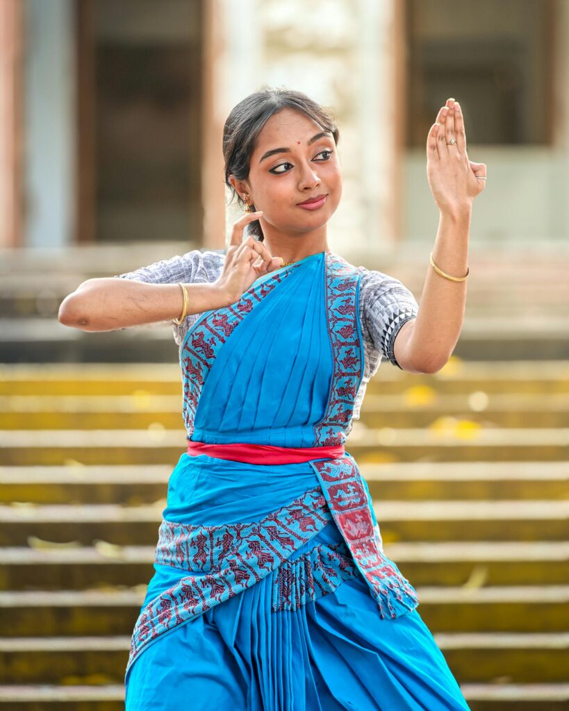 A young woman in traditional attire performing an Indian classical dance outdoors with graceful hand gestures.